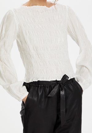 Vrouw draagt een witte gesmokte blouse met pofmouwen en zwarte hoge taillebroek met een gestrikte tailleband.