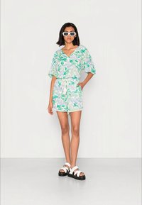 Macacão com padrão floral em tons de verde e azul, apresentando um decote em V, mangas curtas, cintura ajustada e acabamento em renda. Combinado com sandálias brancas.