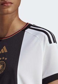 Camiseta deportiva en blanco y negro con cuello rojo, amarillo y negro. Presenta la marca Adidas y el emblema dorado de la federación alemana de fútbol.