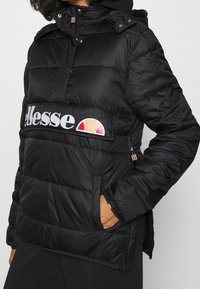 Chaqueta acolchada negra con cuello alto, cierre frontal con cremallera, puños elásticos y bolsillos laterales, que presenta un logo blanco en el pecho.