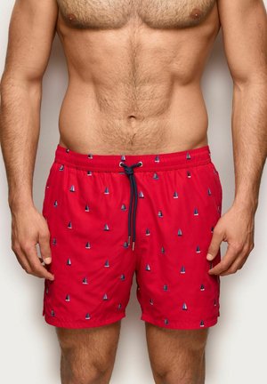 Torso maschile adulto che indossa pantaloncini da bagno rossi con motivi di barche a vela blu e bianche e un cordino nero annodato in vita.