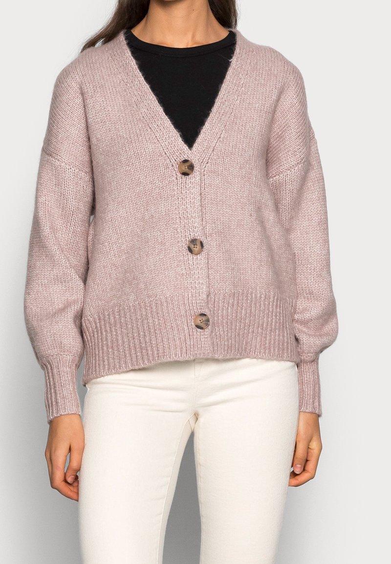 Světle růžový pletený cardigan s výstřihem do V, žebrovaným lemem a třemi velkými knoflíky vpředu; má volné, široké rukávy.
