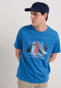 Tricou din bumbac albastru cu un imprimeu grafic cu vele în roșu, albastru, negru și alb. Guler rotund și mâneci scurte.
