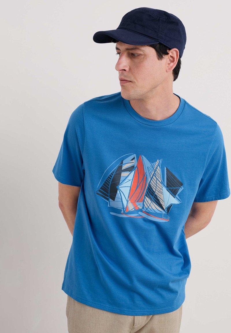 Tricou din bumbac albastru cu un imprimeu grafic cu vele în roșu, albastru, negru și alb. Guler rotund și mâneci scurte.