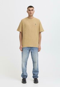 Camiseta de gran tamaño texturizada en color beige con un pequeño logo, combinada con jeans rasgados de color azul claro y zapatos negros, llevada por un modelo de pie.
