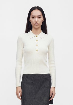 Donna con capelli lunghi e neri che indossa una polo bianca a coste con maniche lunghe, bottoni dorati e una gonna grigia scura con tasche.