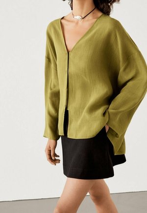 Losse blouse in olijfgroen met een diepe V-hals en knoopsluiting aan de voorkant, gecombineerd met een zwarte rok, met een zachte, gestructureerde stof.