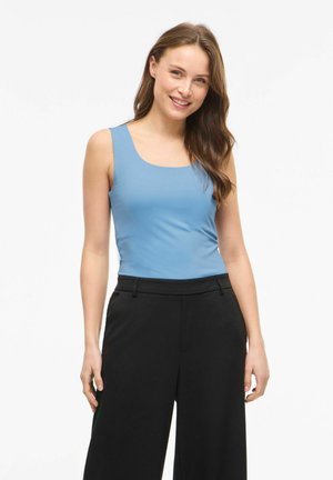 Jeune femme aux longs cheveux bruns portant un haut sans manches bleu et un pantalon noir, souriante sur un fond blanc uni.