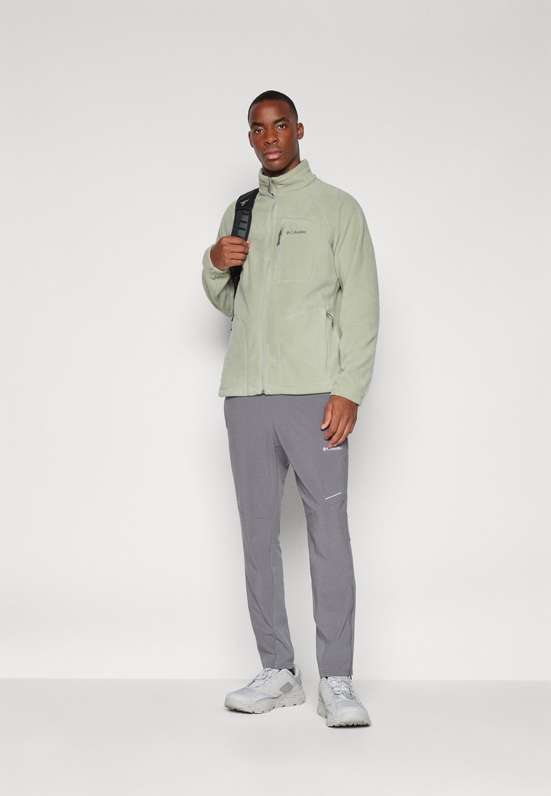 Chaqueta de forro polar verde claro con cremallera, bolsillos laterales y cuello alto, combinada con pantalones ligeros grises y zapatillas blancas.