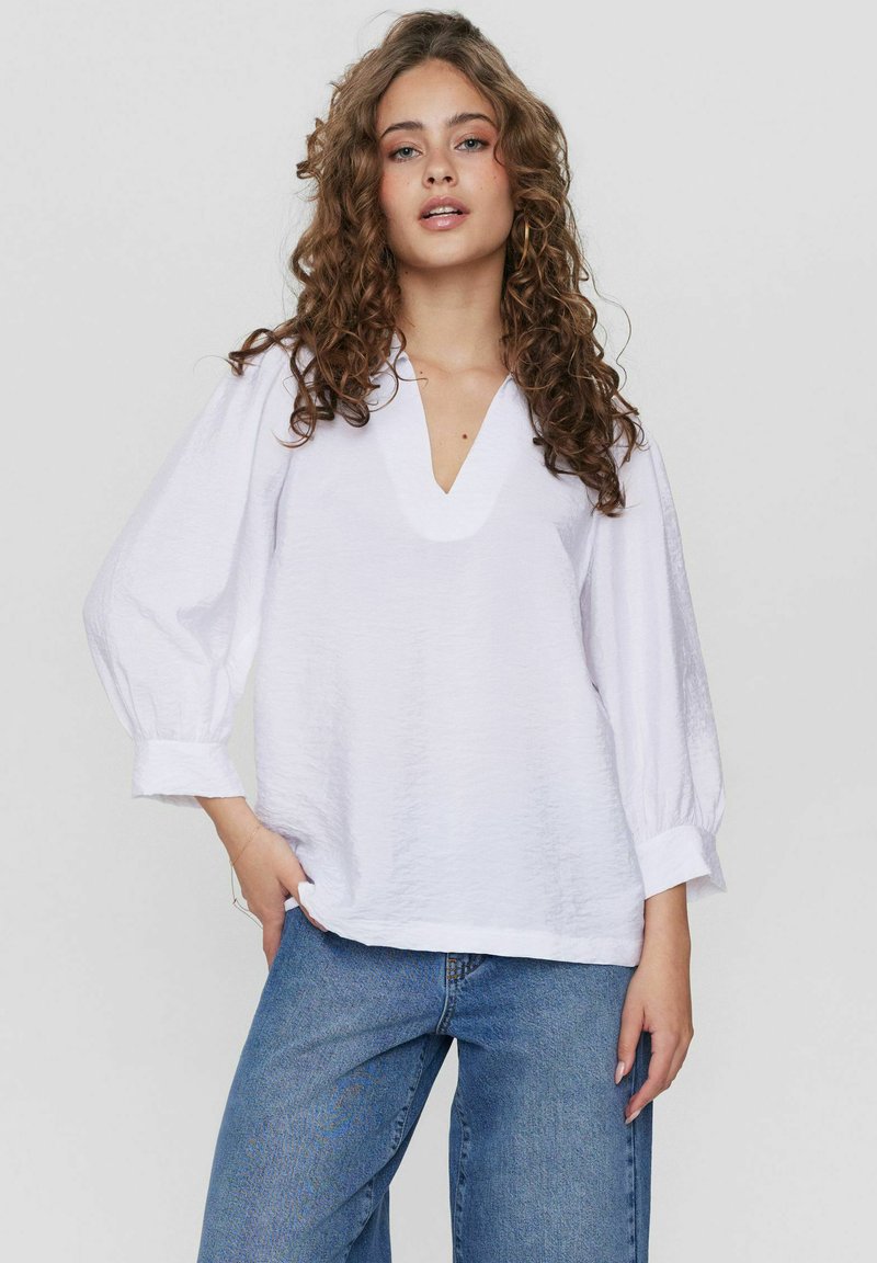 Blusa bianca con texture, scollatura a V e maniche larghe plissettate. Abbinata a jeans blu a gamba larga.