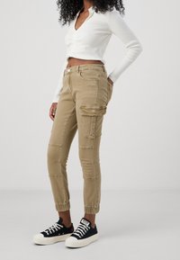 Hnědé cargo kalhoty s bočními kapsami, elastickými manžetami a prošívanými detaily. Spojené s černými plátěnými teniskami s bílými tkaničkami.