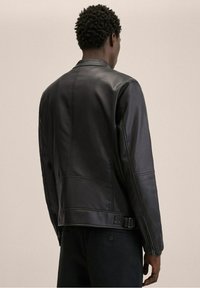 Zwarte leren jas met een gladde textuur, een aansluitend ontwerp en een rits aan de achterkant. Voorzien van op maat gemaakte stiksels en een verstelbare zijriem.