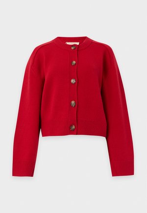 Femme aux longs cheveux noirs portant un cardigan rouge boutonné, une chemise blanche, une ceinture noire, un jean foncé et des bottines noires à talons, debout sur fond blanc.
