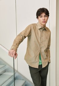 Camisa de botones marrón claro hecha de una tela texturizada, llevada sobre una camiseta verde. La camisa presenta un pequeño parche con un logo y un cuello clásico.
