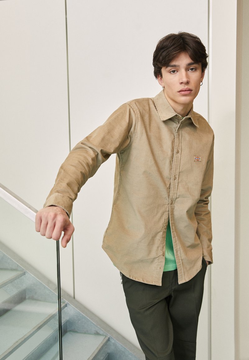 Camisa de botones marrón claro hecha de una tela texturizada, llevada sobre una camiseta verde. La camisa presenta un pequeño parche con un logo y un cuello clásico.