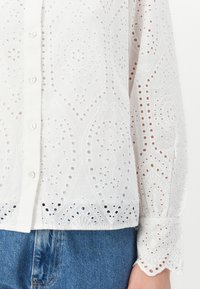 White shirt with intricate cut-out patterns, featuring a button front, long sleeves, and gathered cuffs, paired with blue denim.