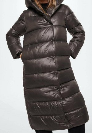 Femme portant un long manteau d'hiver matelassé, brun foncé et brillant, avec un grand col et des poches latérales, debout les mains dans les poches.