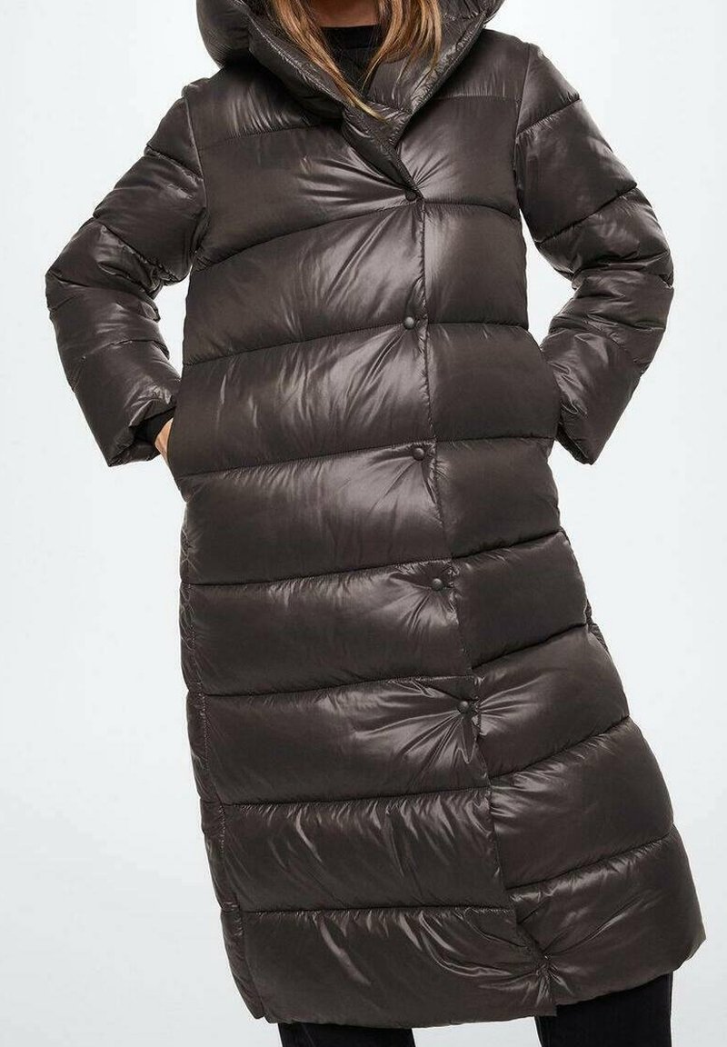 Femme portant un long manteau d'hiver matelassé, brun foncé et brillant, avec un grand col et des poches latérales, debout les mains dans les poches.