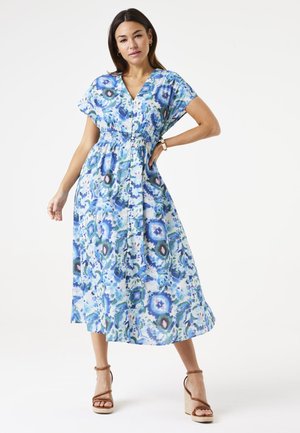 Maxikleid aus blauer und grüner gemusterter Stoff, mit V-Ausschnitt, kurzen Ärmeln und elastischem Bund. Kombiniert mit braunen Riemchenheels.