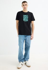 Camiseta de algodón negra con un gráfico en turquesa que dice "NO TE PREOCUPES POR EL FUTURO" en el frente, combinada con jeans de mezclilla azul claro y zapatillas deportivas.