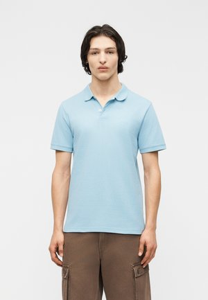 Joven con cabello oscuro que lleva un polo de manga corta azul claro y pantalones cargo marrones, de pie contra un fondo claro y liso.