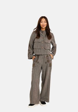 Mujer de pie con chaqueta gris de rayas y pantalones anchos con adornos de cuentas, y zapatos blanco y negro, manos en los bolsillos.