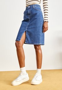 Jeanskjol i mellanblått med slits fram och bältesöljar, i kombination med vita sneakers och ribbade vita strumpor mot en gul bakgrund.