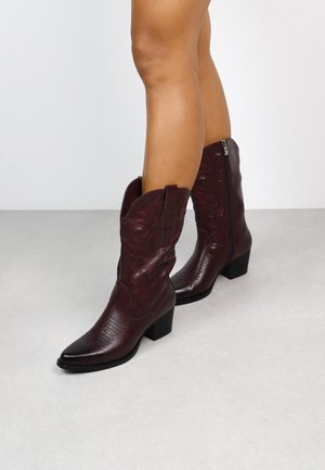 Piernas que llevan botas de vaquero de cuero burdeos oscuro con patrones bordados y tacones anchos, de pie sobre un suelo gris claro.