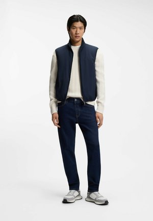 Mężczyzna ubrany w granatową kamizelkę bez rękawów na kremowym swetrze dzianinowym, ciemnoniebieskie dżinsy i białe oraz szare tenisówki, stoi na białym tle.