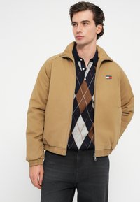 Καφέ bomber jacket με φερμουάρ στο μπροστινό μέρος, ριμπ μανσέτες και μικρό λογότυπο στο στήθος. Φορεμένο πάνω από ένα ναυτικό πουλόβερ με ρόμβους.