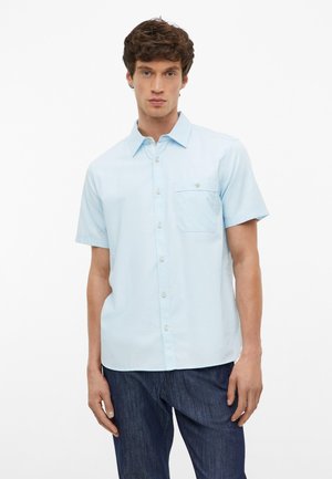 Homme portant une chemise bleu clair à manches courtes boutonnée avec poche poitrine et pantalon bleu foncé, debout devant un fond blanc uni.