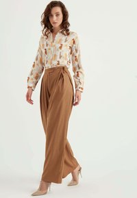 Weiße Bluse mit orangefarbenem Pinselstrichmuster, kombiniert mit braunen, hochgeschnittenen, weiten Hosen und klaren High Heels.