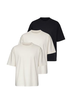 Homme portant un t-shirt beige oversize, un jean bleu clair, des baskets blanches et une montre-bracelet, debout devant un fond blanc uni.