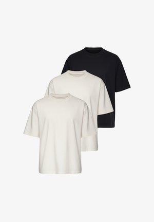 Man draagt een oversized beige T-shirt, lichtblauwe spijkerbroek, witte sneakers en een horloge, staand tegen een effen witte achtergrond.