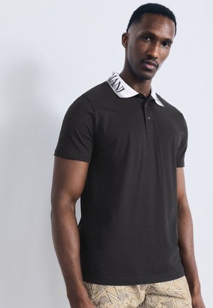 Άνδρας με μαύρο πόλο πουκάμισο με λευκό γιακά κειμένου και μπεζ παντελόνι με σχέδιο, στέκεται μπροστά σε απλό ανοιχτό φόντο.