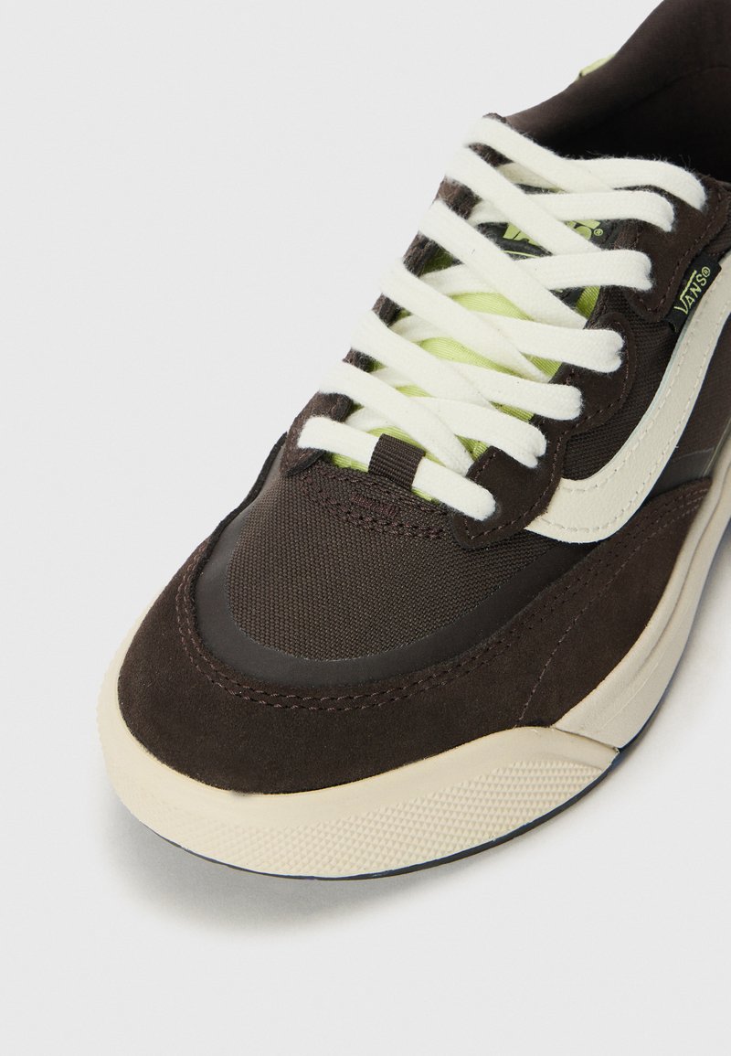 Zapatilla marrón con materiales de ante y tela. Presenta cordones blancos, acentos en verde lima y una suela de goma moldeada con patrones texturizados.