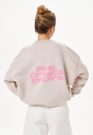 Frau mit Dutt, die einen beigen Sweatshirt mit pinkem Text "Feel. Create. Inspire." auf dem Rücken und weiße Hosen trägt, vom Rücken gezeigt.
