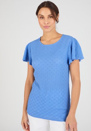 Mujer con blusa azul texturizada de manga corta con mangas con vuelo y pantalones blancos, de pie frente a un fondo claro y liso.