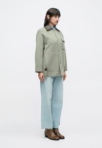 Ženska stoji oblečena v svetlozeleno jakno z gumbi in temnim ovratnikom, svetlo modre široke kavbojke in rjave gležnjarje.