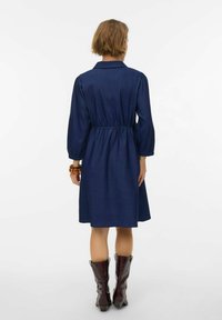 Tmavo plavi traper haljina s uskim pojasom, dugim puf rukavima i ovratnikom, koja ima lepršavu suknju. Glatka textura i dizajn do koljena.