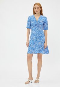 Blauwe bloemenjurk met korte pofdelen, v-hals en uitlopende zoom. Voorzien van een wit bloemenpatroon op een levendige blauwe achtergrond.