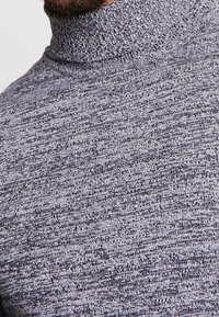 Pull à col roulé en maille gris foncé avec un motif texturé. Le tissu semble doux, présentant des variations subtiles de fils clairs et foncés.