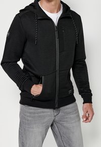 Felpa con cappuccio nera con zip, realizzata in materiale a coste testurizzato, tasche frontali, cappuccio con coulisse e dettaglio con zip laterale. Presenta una toppa con logo all'orlo.