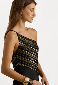 Μαύρο φόρεμα με σχέδιο ενός ώμου διακοσμημένο με οριζόντιες χρυσές και μαύρες πούλιες. Περιλαμβάνει μια χρυσή βραχιόλι ως αξεσουάρ.