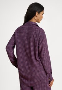 Langermet lilla skjorte med et geometrisk mønster i rødt og blått. Den har krage og knappede mansjetter.