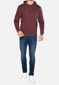 Homme portant un sweat à capuche bordeaux avec une poche avant, un jean slim bleu foncé et des baskets blanches, debout devant un fond uni.
