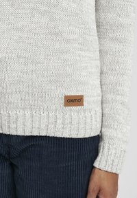 Ljusgrått stickat tröja med ribbad nederkant, med en brun läderetikett med "oxmo." Mörk marinblå kordbyxor är delvis synliga.