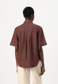 Chemise en lin marron à manches courtes avec un pli au dos et un col, présentant un ourlet arrondi. Le tissu a une finition texturée.