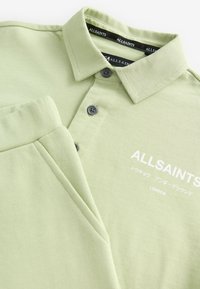 Grøn polo fra AllSaints med korte ærmer, ribbet krave, to-knaps stolpelukning, broderet logo og glat bomuldstekstur.