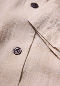 Tela de camisa de lino color tostado con una superficie texturizada, que presenta botones marrón oscuro y un cuello doblado, resaltando los detalles de costura.